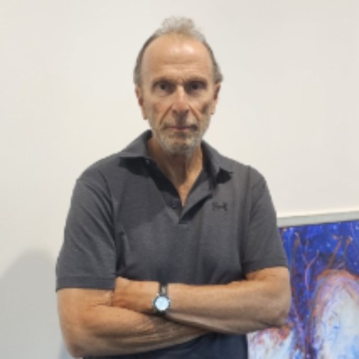 A man with gray hair stands with arms crossed next to colorful paintings leaning against a white wall in a gallery setting.