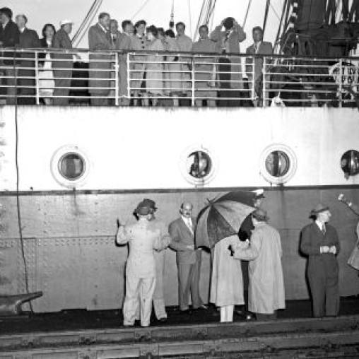 Black and white photo of a ship with people on the upper deck and several individuals on the dock, some holding an umbrella, as they interact near the ship's side.