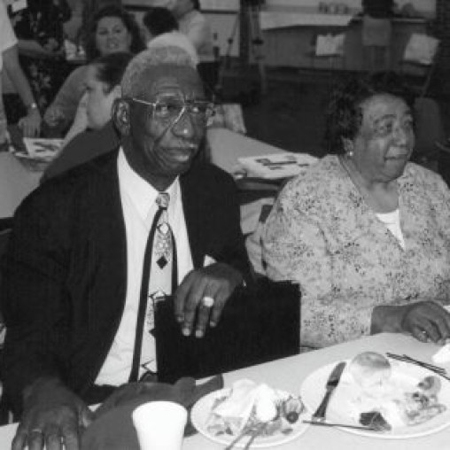 A black and white image of an elderly man in a suit and an elderly woman in a patterned blouse sitting at a table with plates of food.