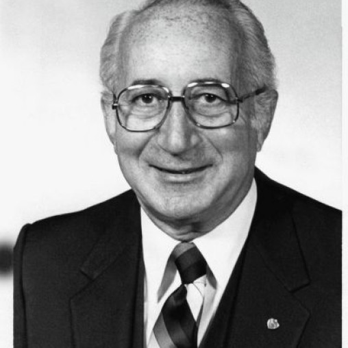 Black and white portrait of an older man with glasses, smiling, wearing a dark suit with a striped tie, and a white shirt.