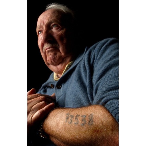 An elderly man with a serious expression, wearing a blue sweater, rests his arms on his lap, revealing a tattoo on his forearm that reads "7538." The background is dark.
