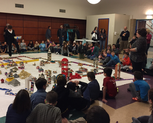 A room full of children and adults with a village in the middle built out of legos