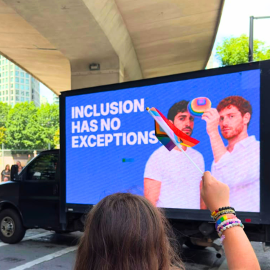 A person holding a small rainbow flag stands in front of a digital billboard displaying two men and the text "INCLUSION HAS NO EXCEPTIONS." The scene is outdoors under a bridge.