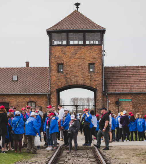 A group of people wearing blue jackets and red hats gathered in front of the entrance gate of Auschwitz, with a watchtower visible above.