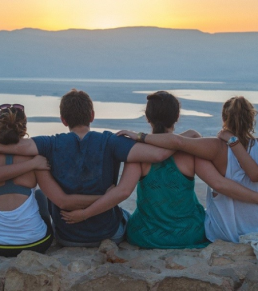 A group of four people sitting on a rocky ledge, embracing each other while watching a sunset over a body of water.