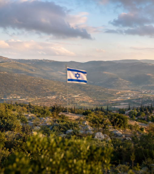 An Israeli flag with a blue Star of David is flying on a flagpole, set against a scenic landscape of rolling hills and cloudy skies.