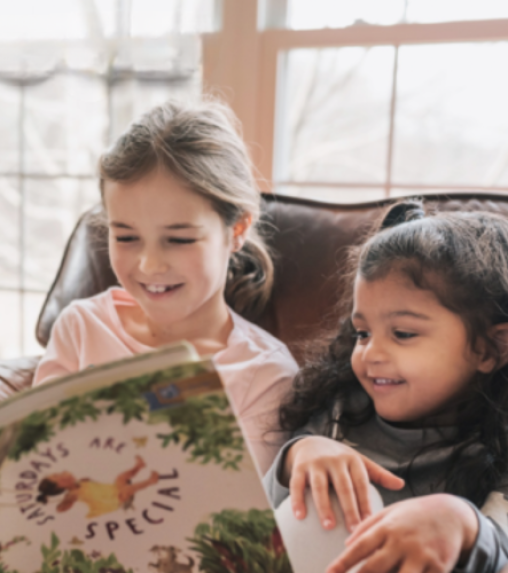 Two young girls sitting on a couch, smiling and reading a book titled "Saturdays are Special." Natural light from a window illuminates the scene.