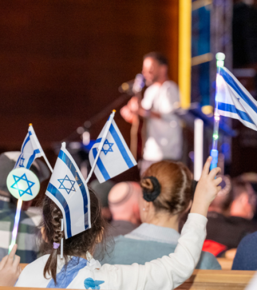 A child holding an Israeli flag raises her hand in a crowd, while a musician performs on stage in the background.