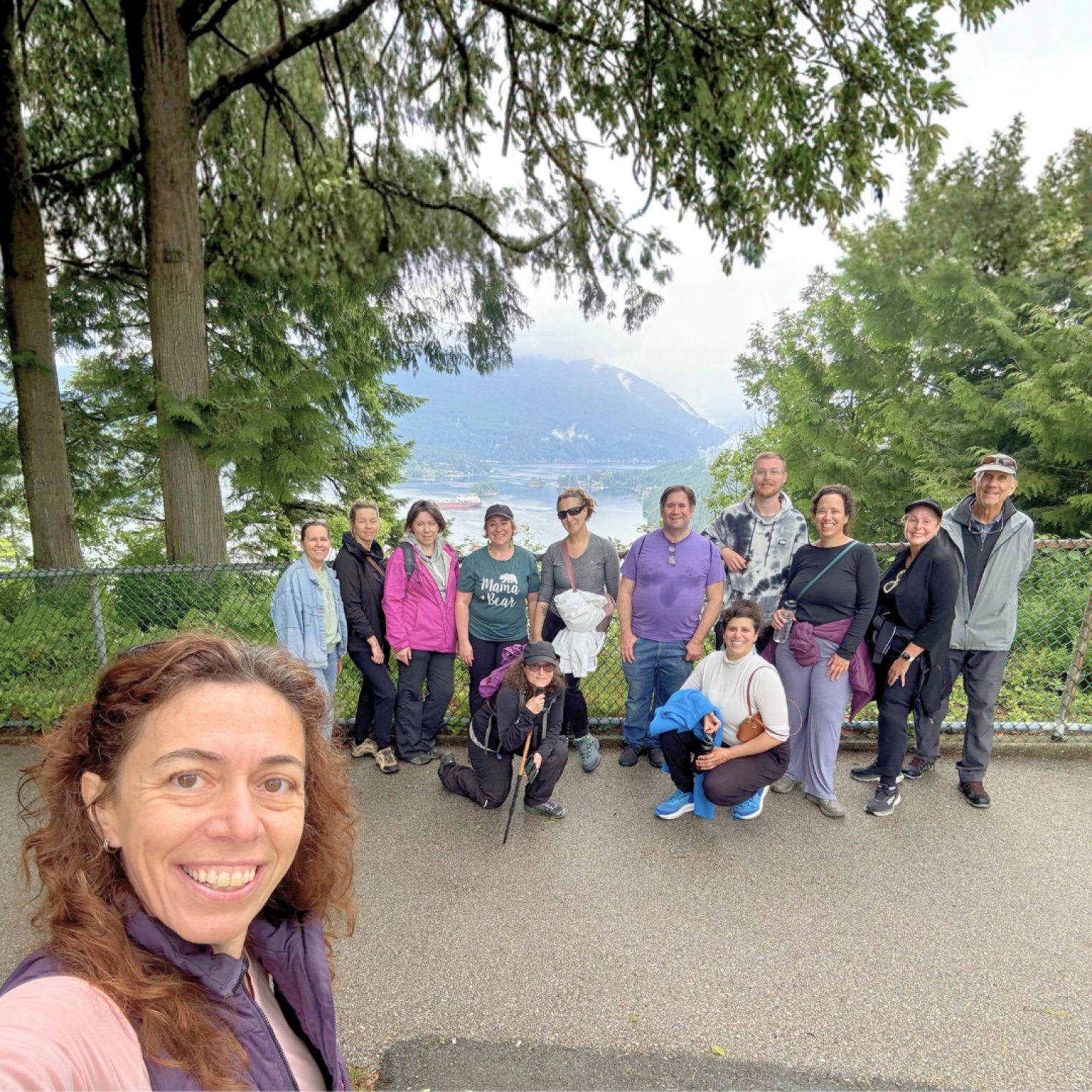 A group of 14 people posing for a photo on a path surrounded by trees, with mountains and a body of water in the background.