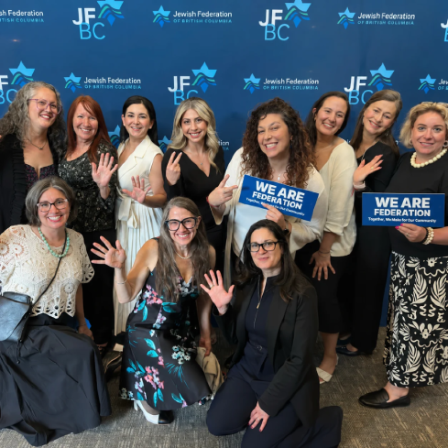 A group of twelve women posing in front of a blue backdrop with "Jewish Federation of British Columbia" text, holding signs that say "WE ARE FEDERATION."