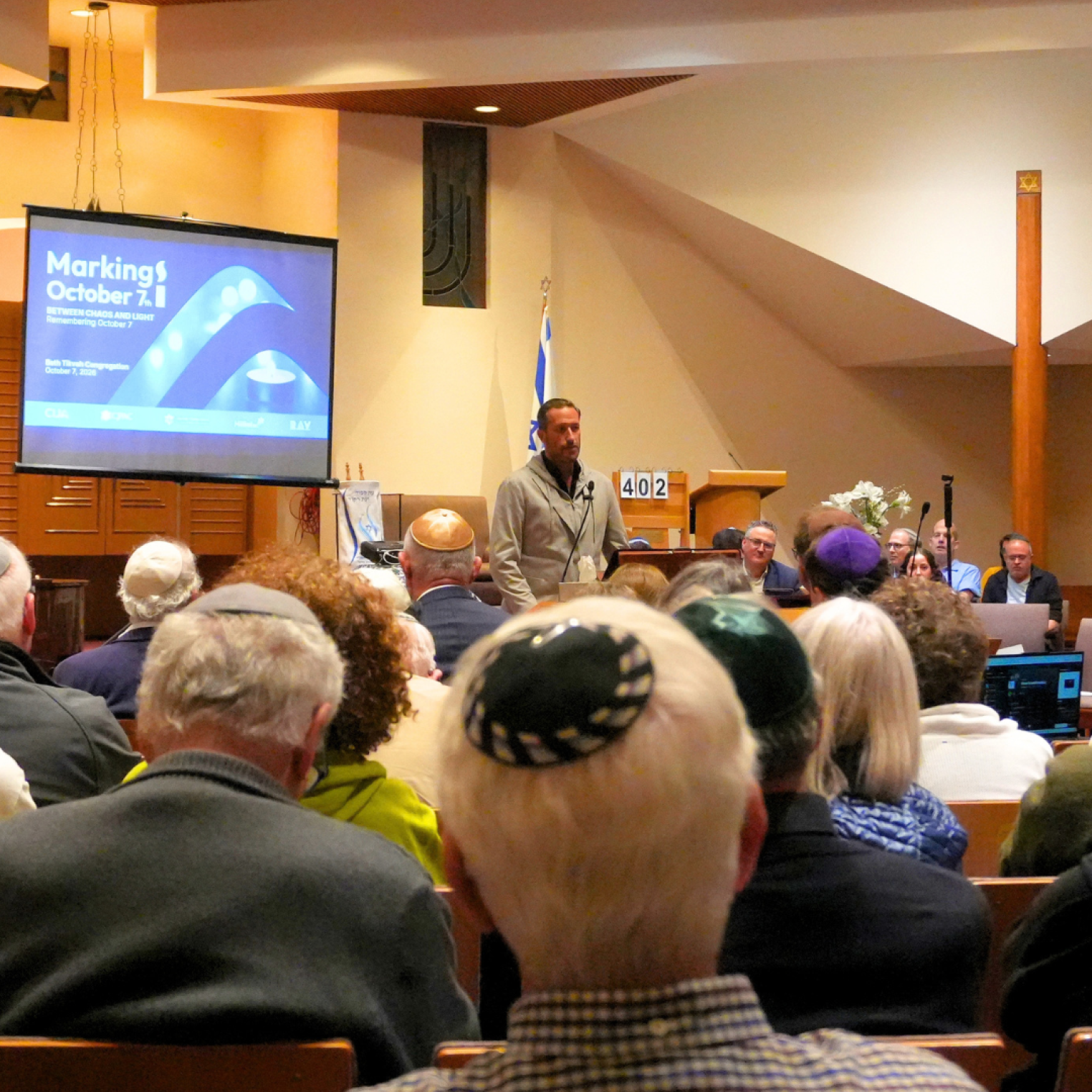 A man speaking at a podium in a synagogue, with a screen displaying "Marking October 7" behind him; attendees wearing kippahs are seated in front.