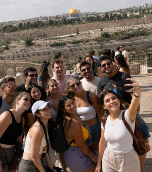 A group of diverse young adults posing for a selfie with the Dome of the Rock visible in the background, surrounded by a scenic landscape.