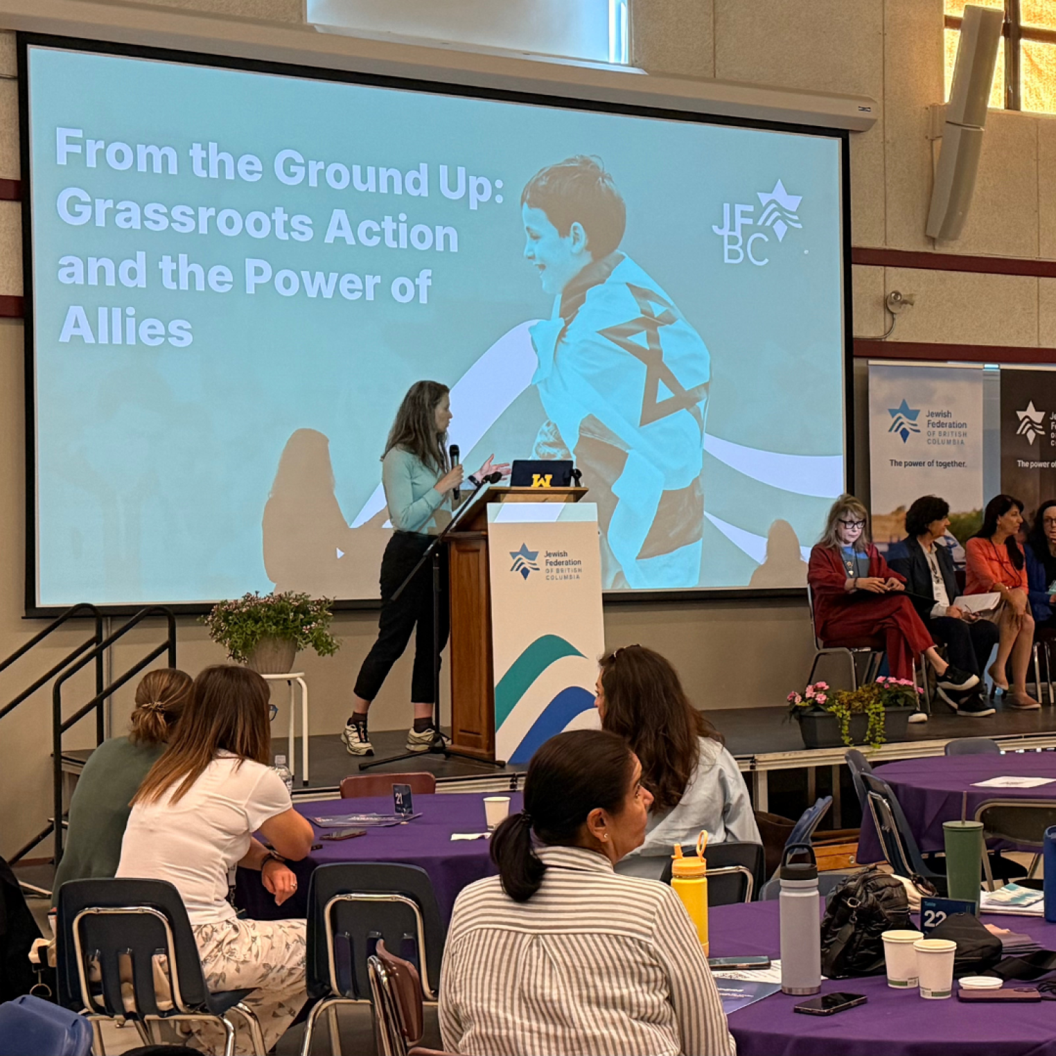 A woman speaking at a podium with a presentation slide reading "From the Ground Up: Grassroots Action and the Power of Allies," while an audience listens.