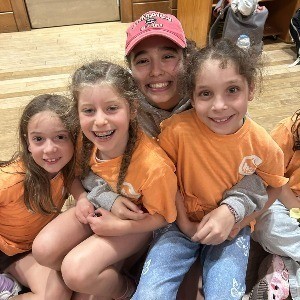 Four children sitting together on a wooden floor, all wearing orange t-shirts. One girl in a pink cap is smiling widely, while the others are also smiling and posing closely.