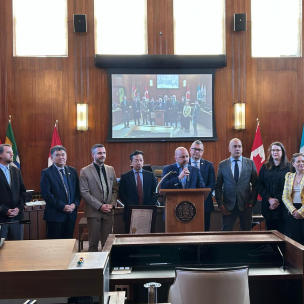 A group of eight individuals stands in a government chamber, with a man speaking at a podium. A screen behind displays a larger group photo. Flags are visible on the walls.