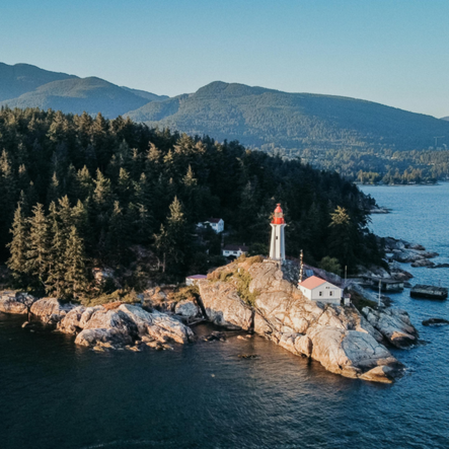 Aerial view of a lighthouse with a red top, situated on rocky land surrounded by trees and water, with mountains in the background.
