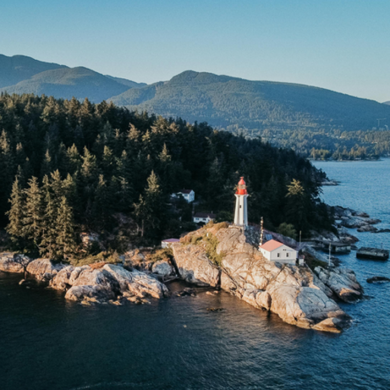 Aerial view of a lighthouse with a red top, situated on rocky land surrounded by trees and water, with mountains in the background.