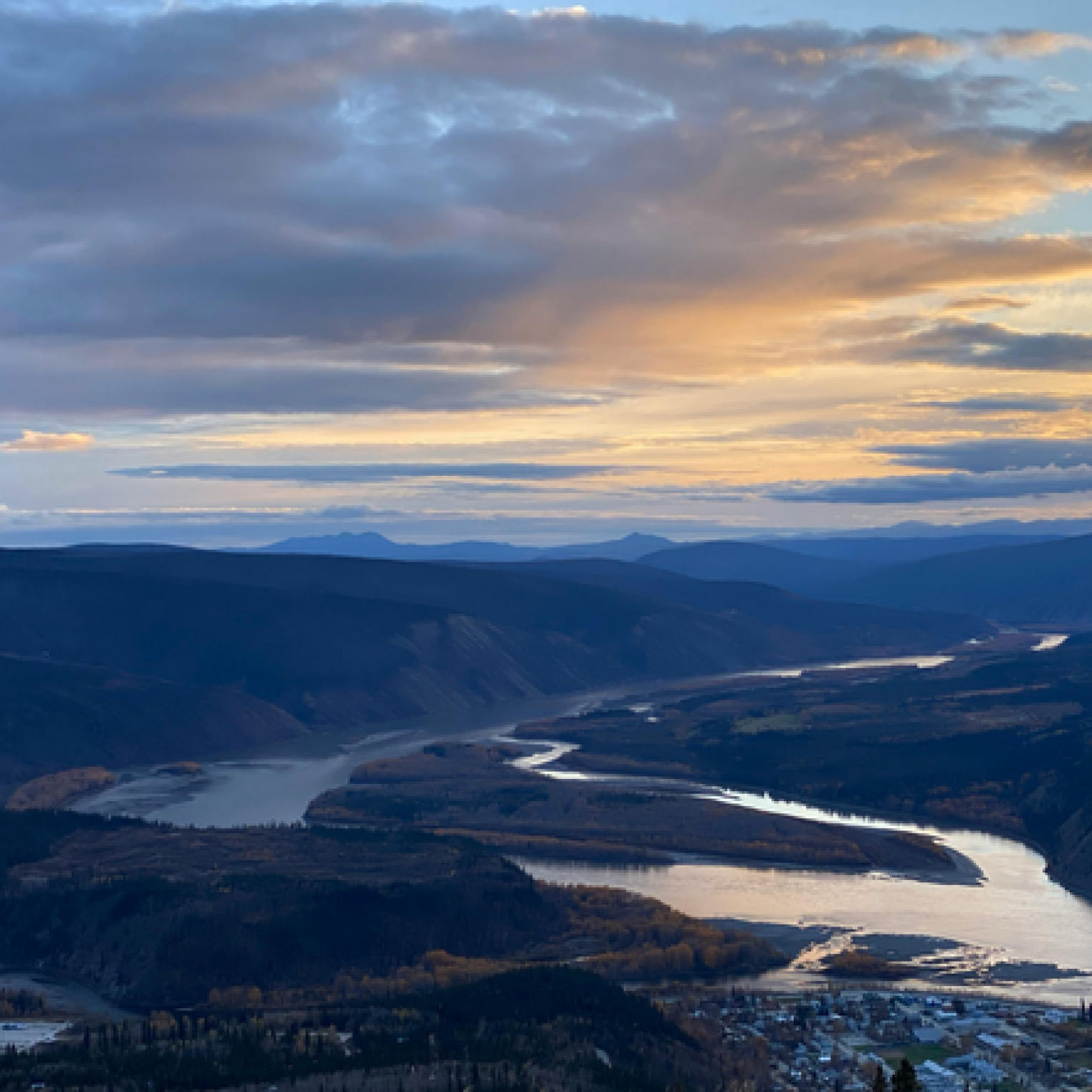 A panoramic view of a winding river surrounded by mountains under a cloudy sky at sunset, with a small town visible in the foreground.