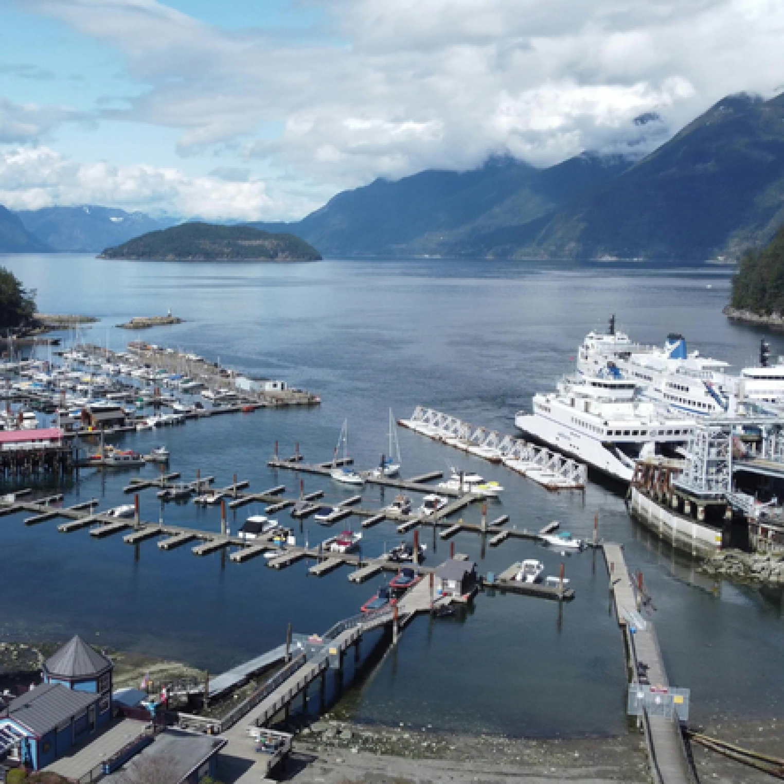 Aerial view of a marina with numerous boats and a large ferry docked, surrounded by mountains and a calm body of water under a partly cloudy sky.
