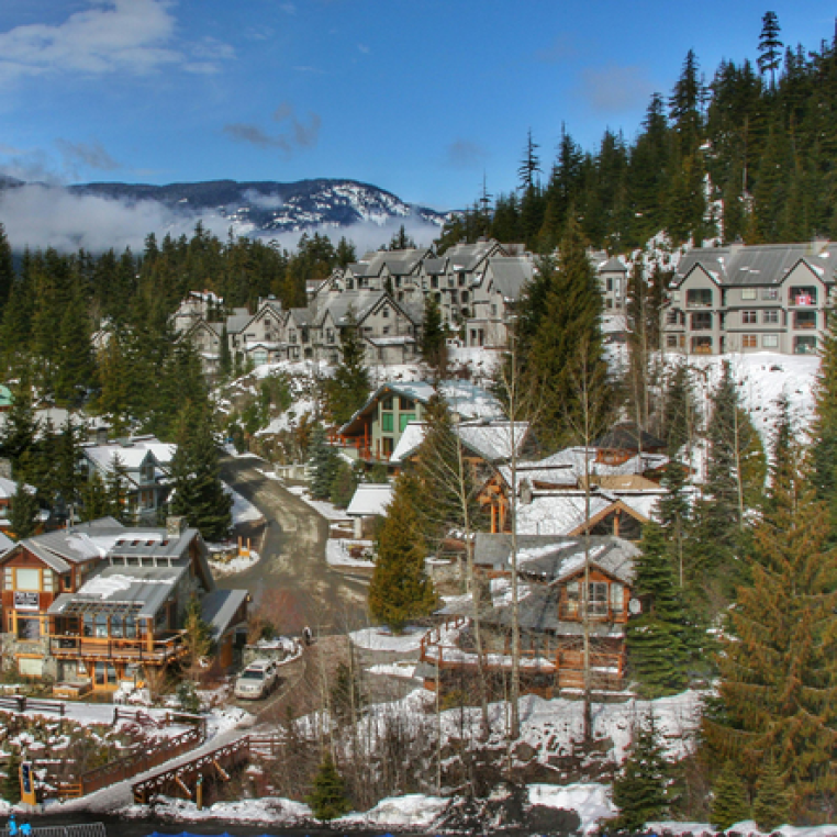 A scenic view of a snowy mountain village with wooden and stone houses, surrounded by evergreen trees and mountains in the background.