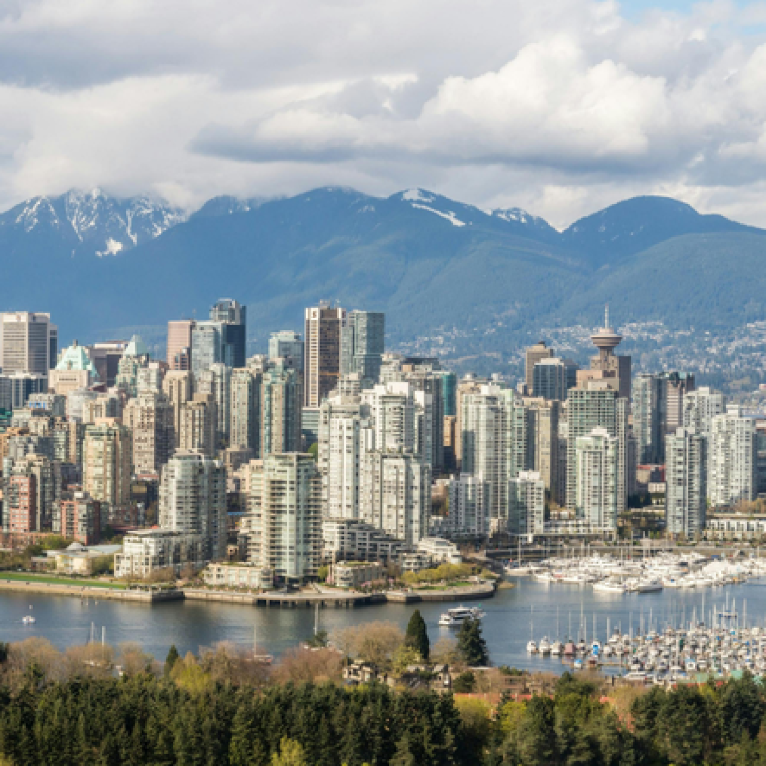 A panoramic view of Vancouver's skyline featuring tall buildings, a marina, and mountains in the background under a partly cloudy sky.