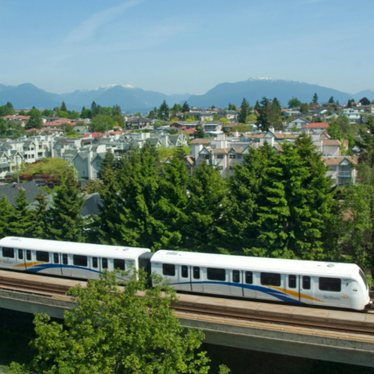 A light rail train traveling on an elevated track, surrounded by green trees and residential buildings, with mountains in the background.
