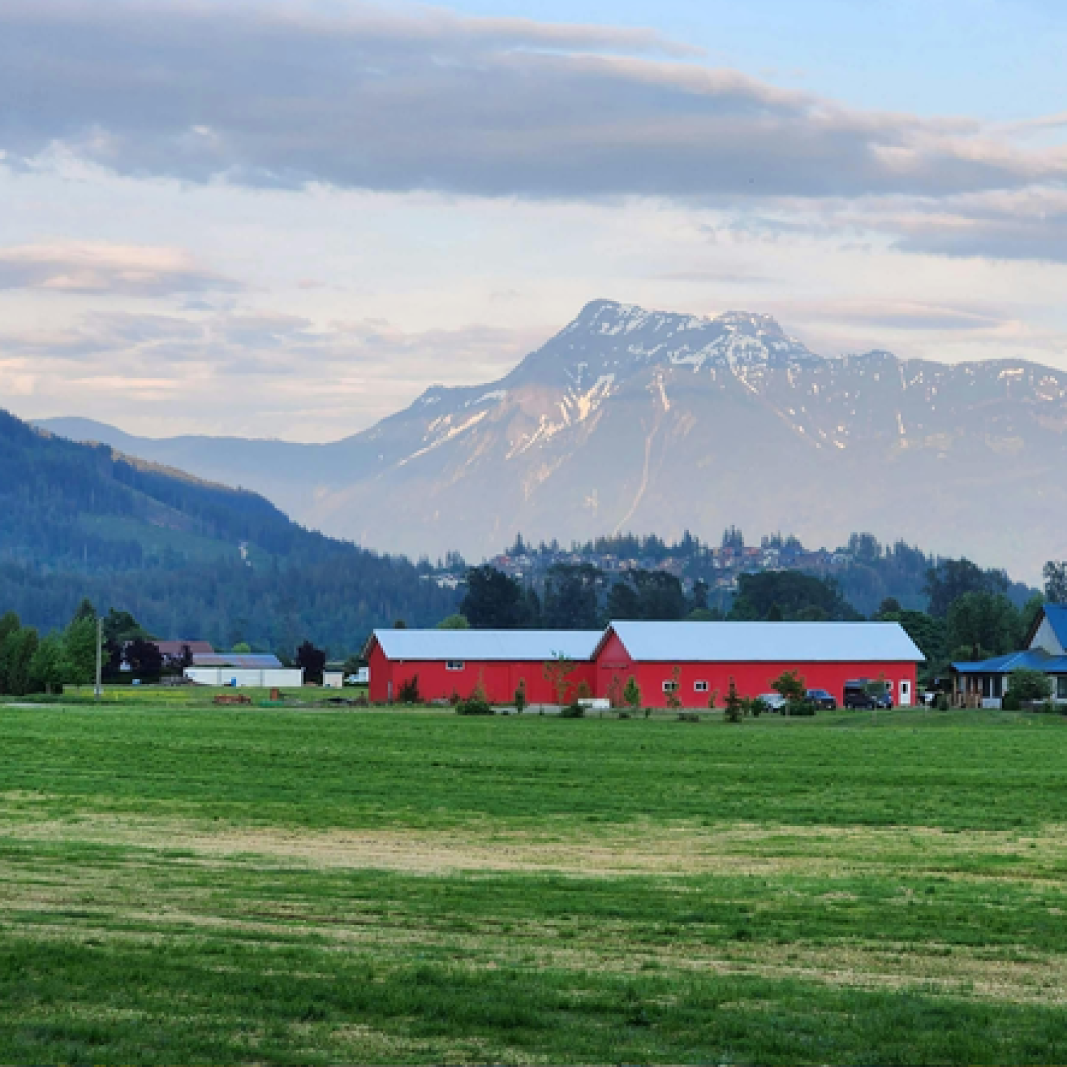 A scenic view of a green field with a red barn and a blue-roofed building, set against a backdrop of snow-capped mountains under a cloudy sky.