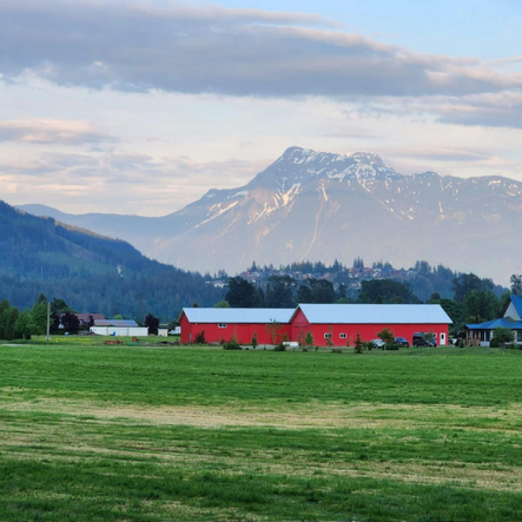 A scenic view of a green field with a red barn and a blue-roofed building, set against a backdrop of snow-capped mountains under a cloudy sky.