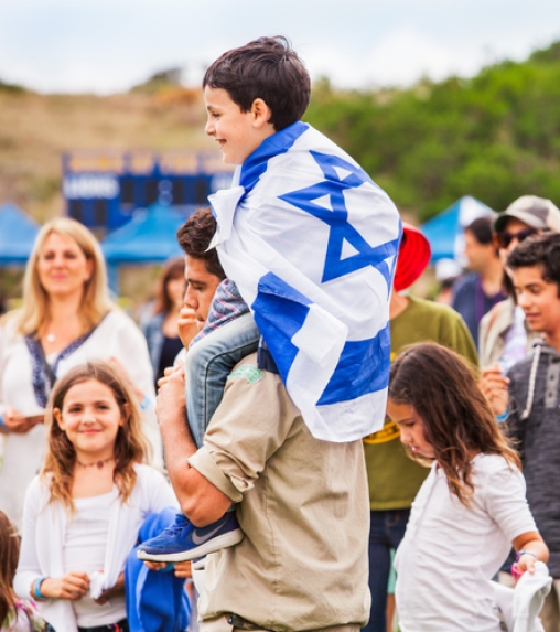 A boy wearing an Israeli flag is sitting on the shoulders of a young man, surrounded by a diverse group of children and adults at an outdoor event.