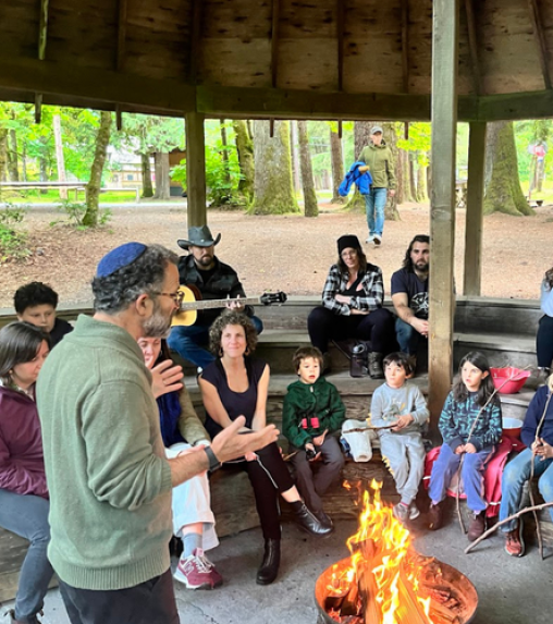 A group of adults and children gathered around a fire in a gazebo, with a man speaking and another playing guitar. Trees are visible in the background.
