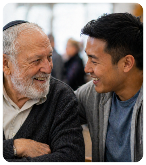Two men smiling at each other, one elderly with a kippah and a beard, and the other younger with short hair, seated indoors.