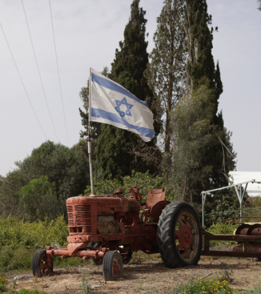 An old red tractor with a blue and white Israeli flag attached, set against a backdrop of tall trees and greenery.