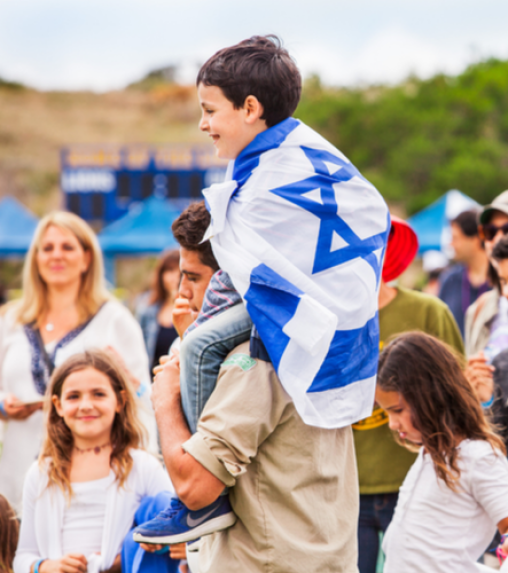 A boy wearing an Israeli flag is sitting on a man's shoulders, surrounded by a diverse group of people at an outdoor event.