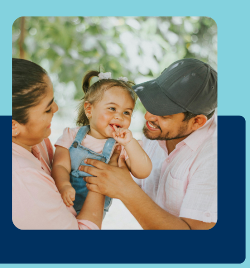 A woman and a man smiling at a toddler in overalls, who is playfully touching her lips, surrounded by greenery.