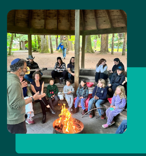 A group of children and adults gathered around a fire in a gazebo, with a man speaking animatedly and others listening. Some participants are holding sticks over the fire.