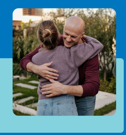 A man and a woman embrace each other in a garden setting, surrounded by greenery and a pathway. The woman has her hair in a bun and wears a purple sweater, while the man is bald and dressed in a maroo