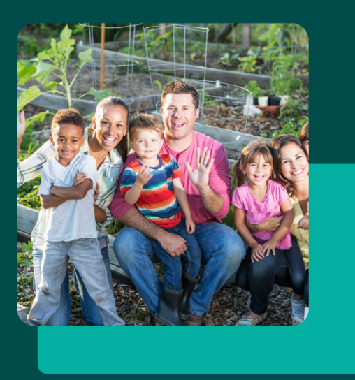 A diverse group of six people, including three children, smiling and waving while sitting in a garden with plants in the background.