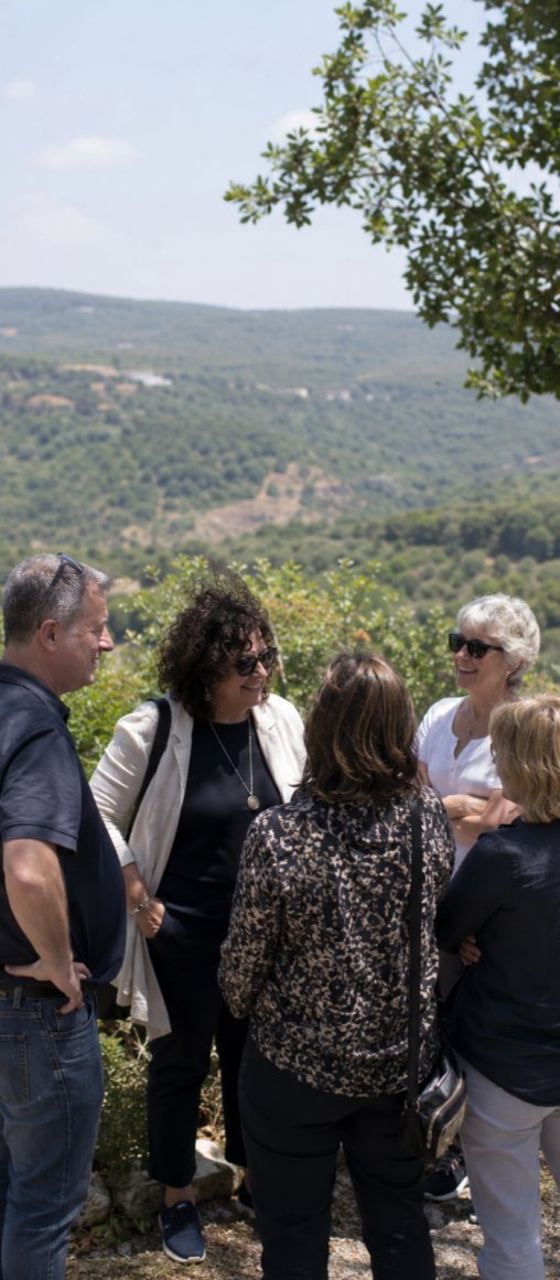 A group of five adults, including three women and two men, are engaged in conversation outdoors with a scenic view of green hills in the background.