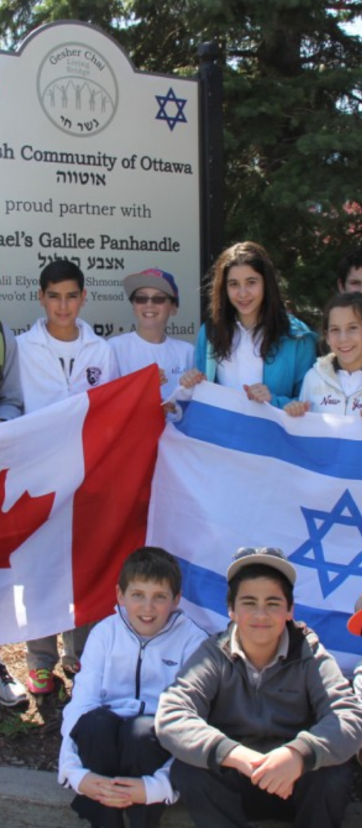 A group of children holding a Canadian flag and an Israeli flag stands in front of a sign that reads "Jewish Community of Ottawa."