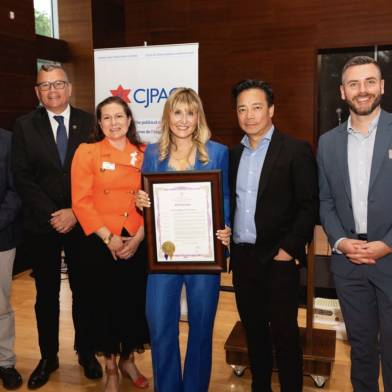 A group of six people poses for a photo, with a woman in a blue suit holding a framed certificate in the center. They are in a room with wooden walls and a banner in the background that reads "CJPAC."