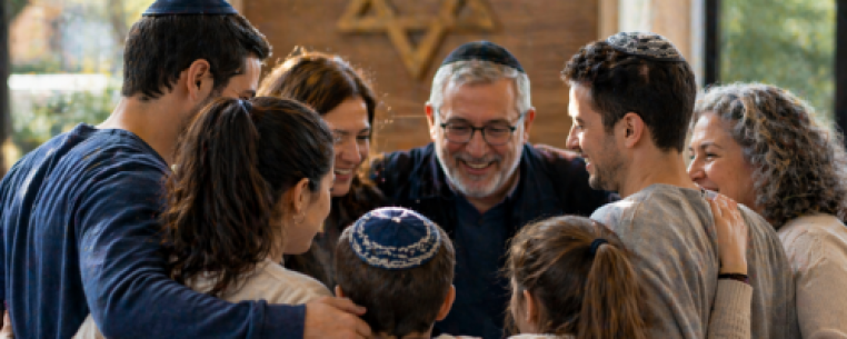 A group of eight people, wearing kippahs, standing in a circle with arms around each other, smiling together in a warmly lit room with a Star of David in the background.