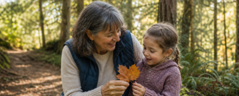 A woman and a young girl smiling while holding a maple leaf, surrounded by trees in a forest setting.