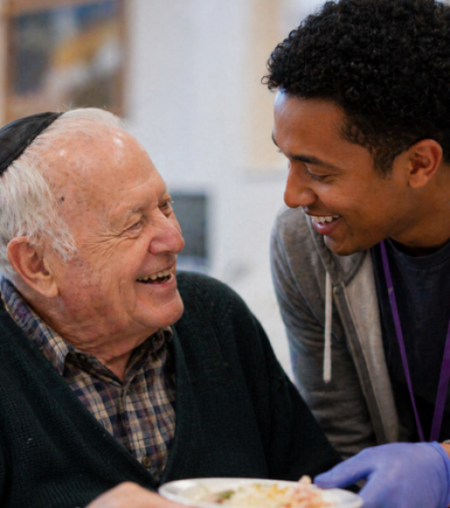 An elderly man wearing a kippah smiles at a younger man, who is wearing gloves and a gray hoodie, while holding a bowl of food.