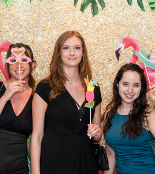 Three women pose in front of a glittery gold backdrop decorated with flamingos, holding playful photo props: a pink flamingo mask and a colorful parrot.