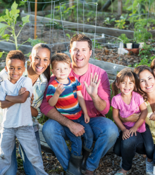 A group of six adults and children smiling and posing together in a garden, with plants growing in the background.