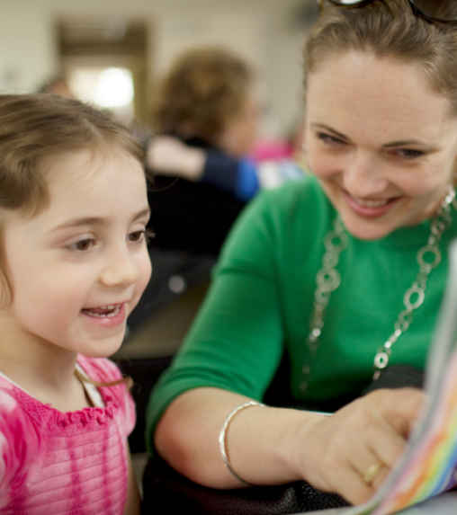 A woman in a green shirt and a girl in a pink shirt are sitting together, looking at a colorful page with smiles.