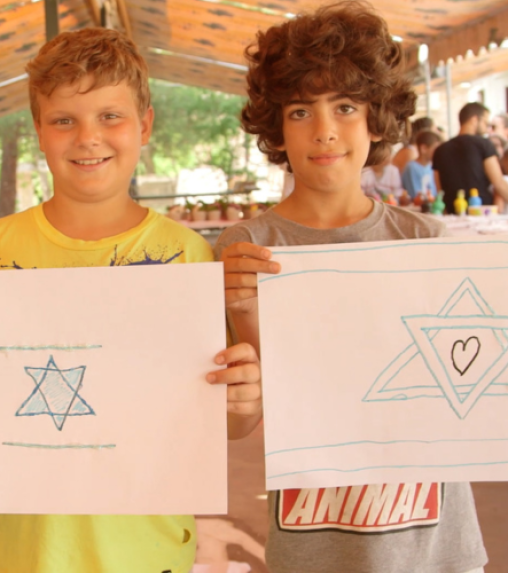 Two boys stand side by side, each holding a piece of paper with a drawing of a Star of David. The boy on the left has a yellow shirt, and the boy on the right wears a gray shirt with the word "ANIMAL.