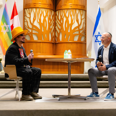 A man in a bright yellow hat and black outfit sits with a microphone, facing another man in a blue jacket at a round table, with a wooden backdrop featuring a tree design and flags of Canada, Israel,