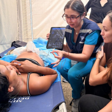 A healthcare professional in scrubs uses a tablet to show an ultrasound image to a woman lying on a medical bed, while another woman observes.