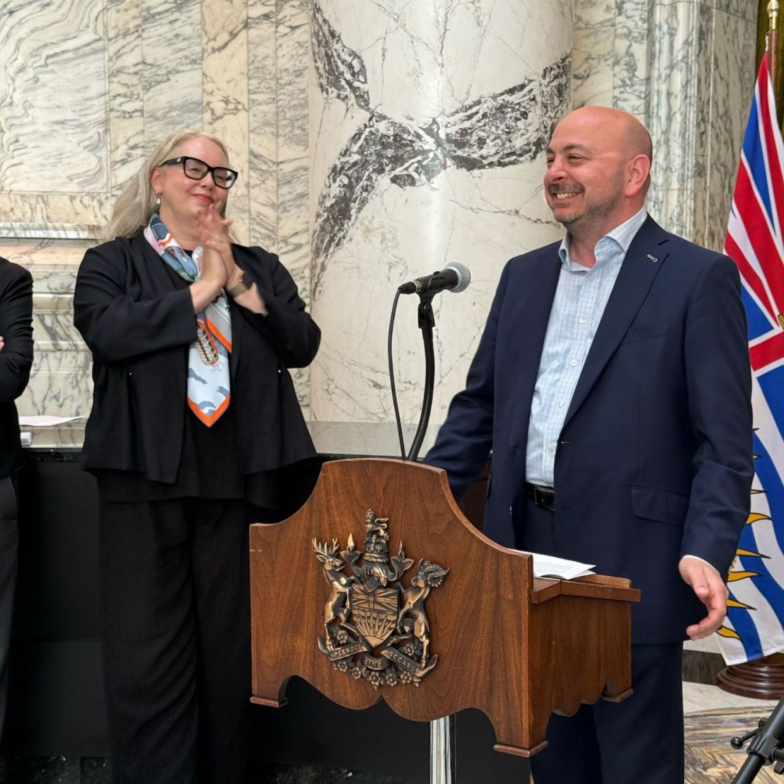 A man in a suit speaks at a podium with a coat of arms, while a woman in glasses and a scarf claps beside him. A microphone is positioned on the podium, and a flag is visible in the background.